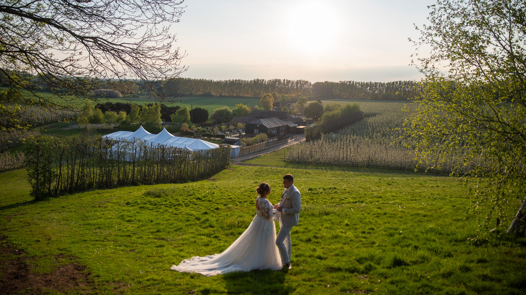 A bride and groom in the top field at The BarnYard at golden hour, the Arboretum and the orchards spread out below them