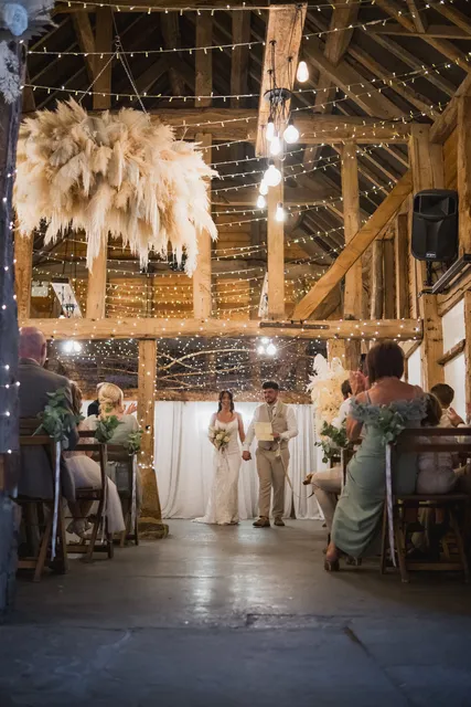 A couple at the altar in the Apple Barn, pampas grass and fairy lights overhead