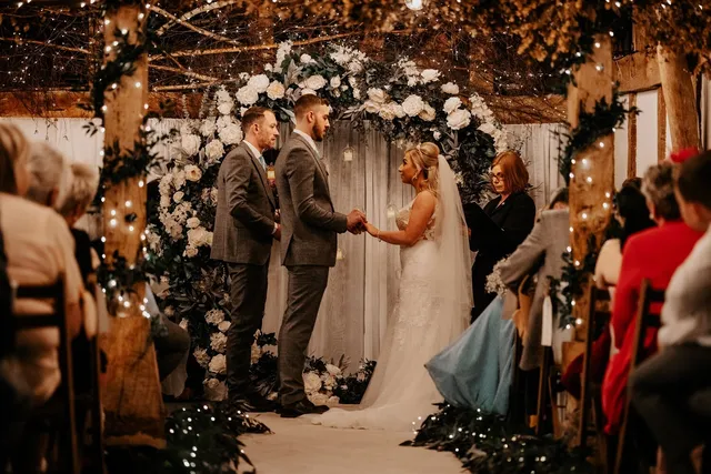 A couple hold hands at a white floral arch during their Apple Barn ceremony