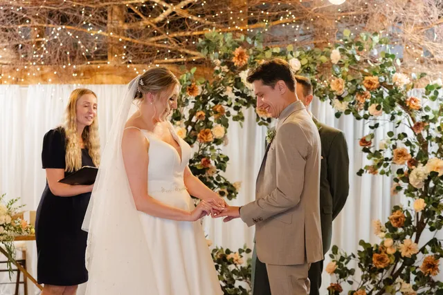 A couple exchange rings beneath a peach and greenery arch in the Apple Barn