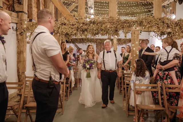 A bride walks the aisle on her father's arm past seated guests in the Apple Barn