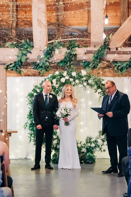 A couple and their celebrant beneath a greenery and white rose arch in the Apple Barn