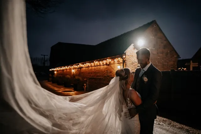 A couple outside the lit barns at night, the bride's veil trailing