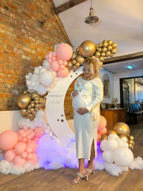 A mum-to-be beside a pink and gold balloon arch at her baby shower