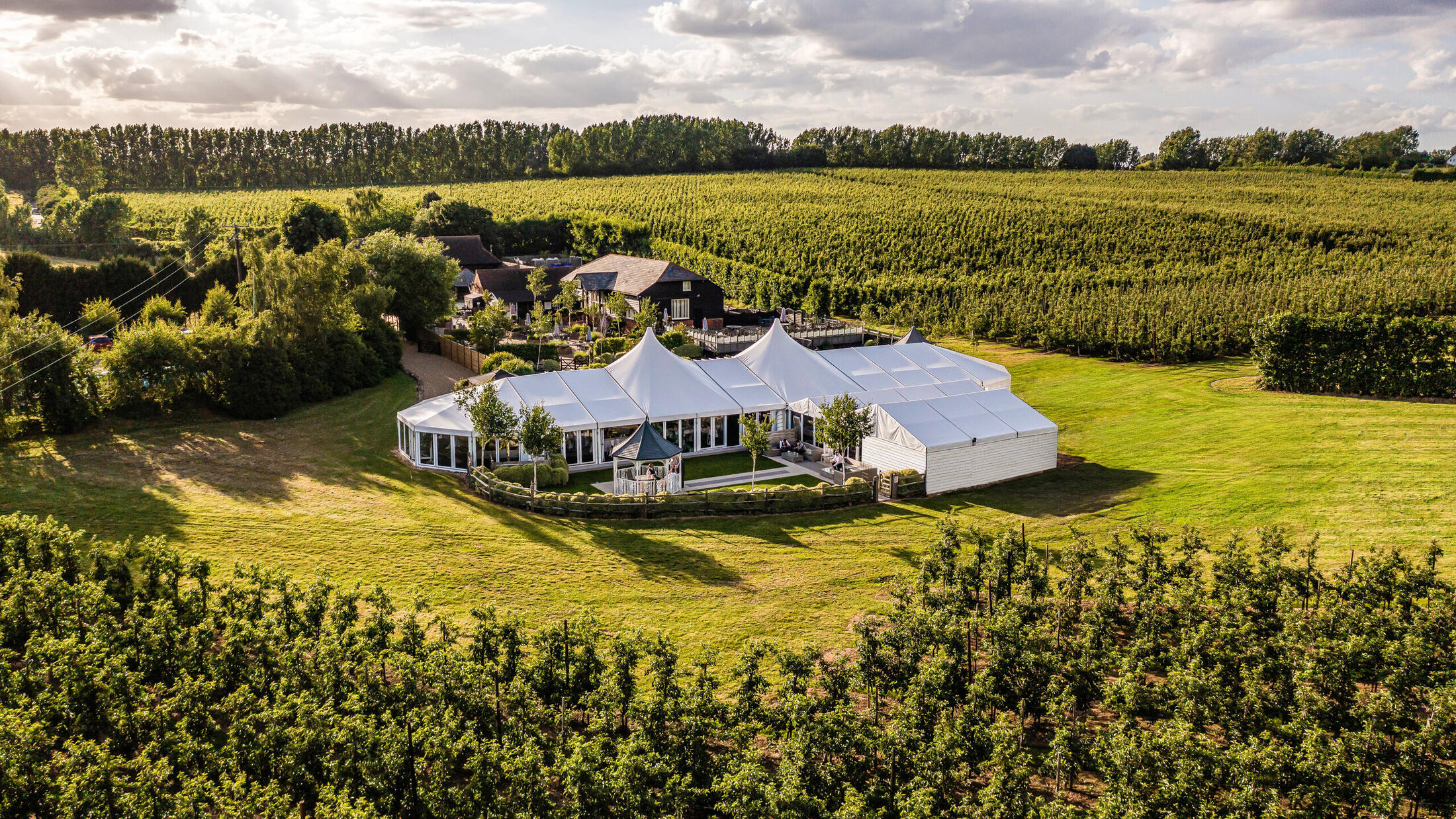 The BarnYard wedding venue from above, glass pavilion surrounded by Kent orchards at golden hour
