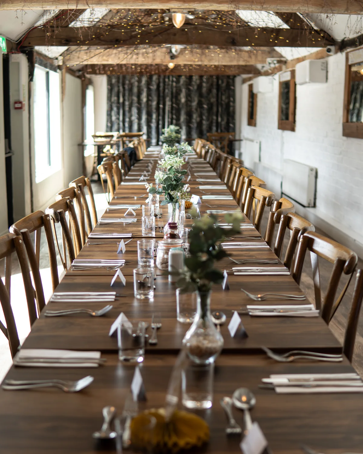 A long banquet table laid for an event beneath the fairy-lit rafters at The BarnYard in Kent