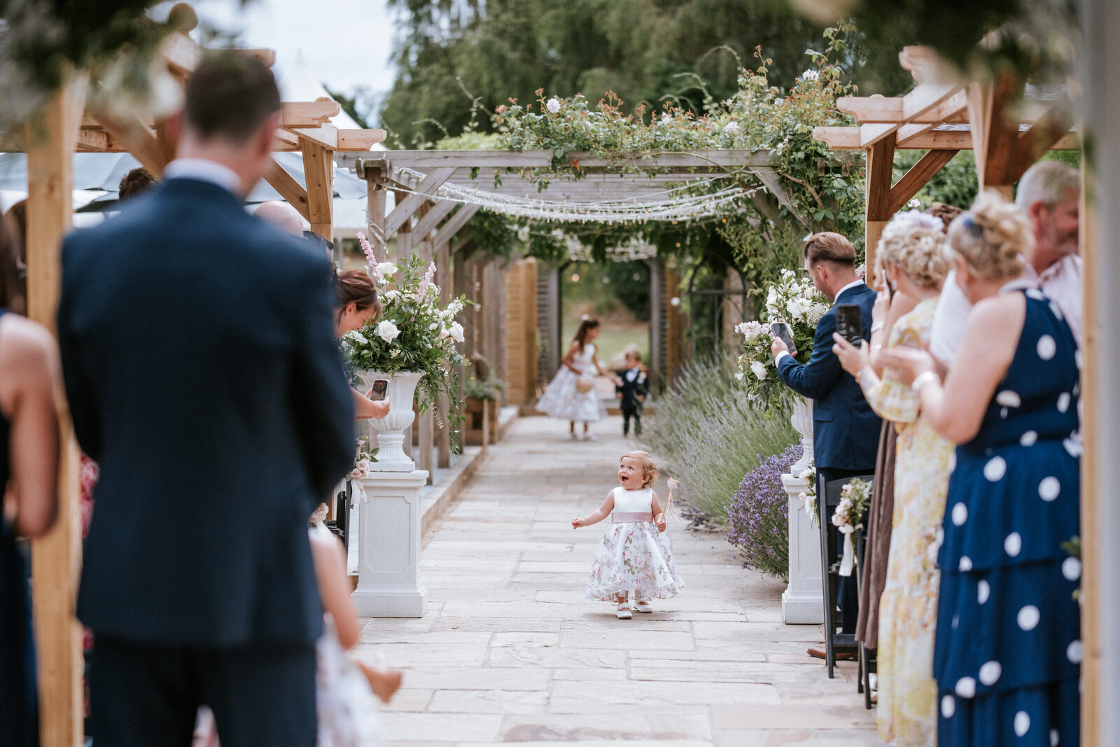 A flower girl running down the aisle beneath the Lavender Walk pergola at The BarnYard