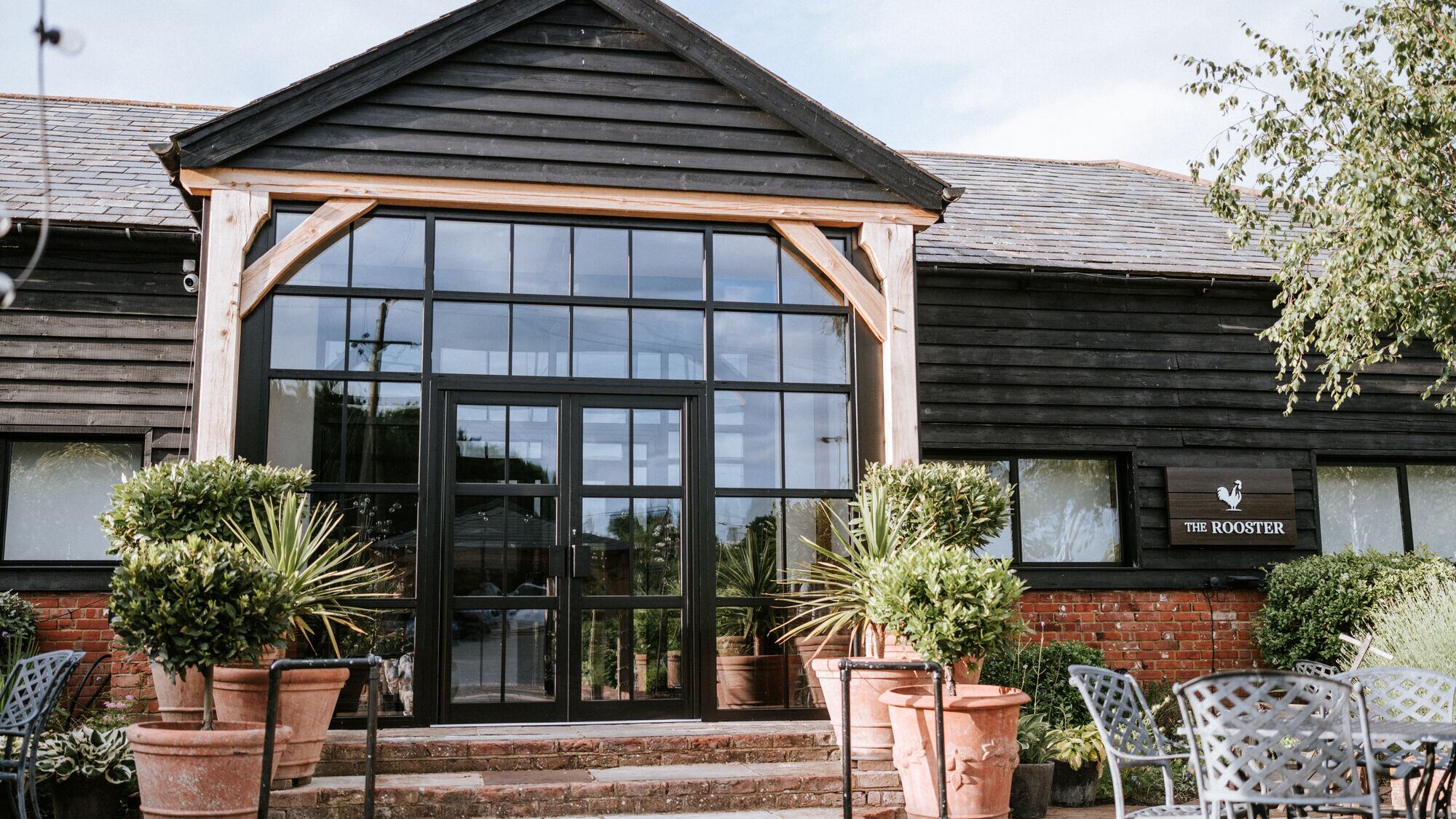The glazed A-frame entrance to The Rooster at The BarnYard, framed by potted olive trees