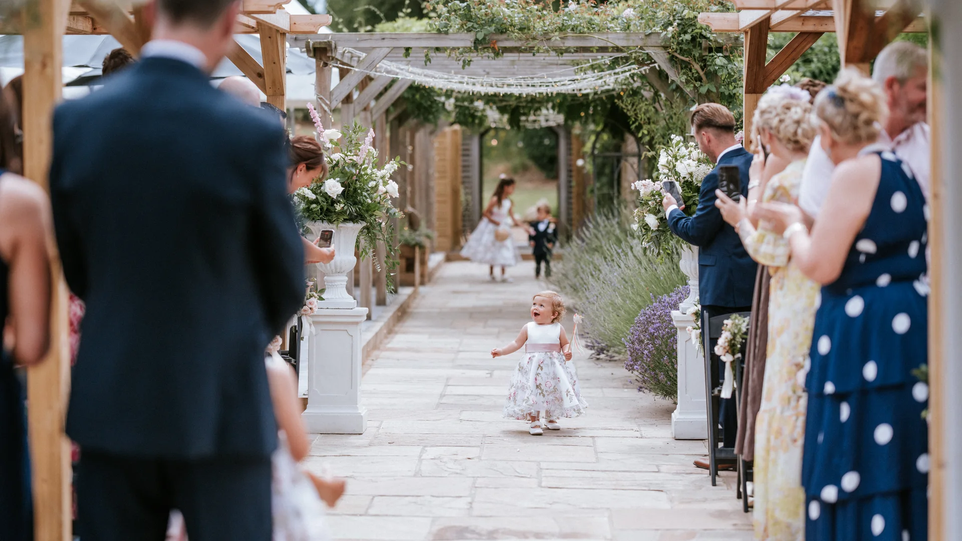 A flower girl running down the aisle beneath the Lavender Walk pergola at The BarnYard