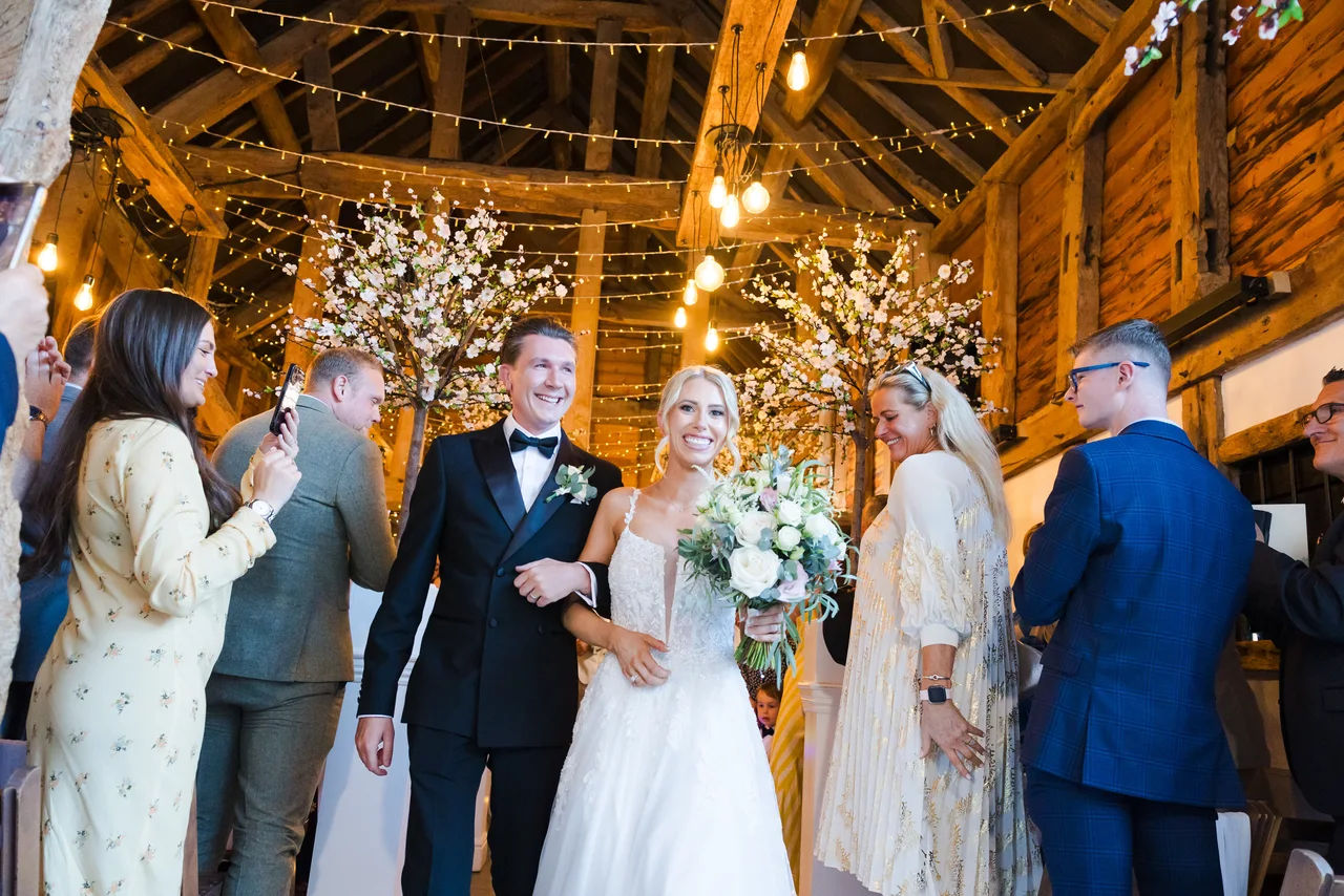 A newly married couple walking back down the aisle in the fairy-lit Apple Barn at The BarnYard