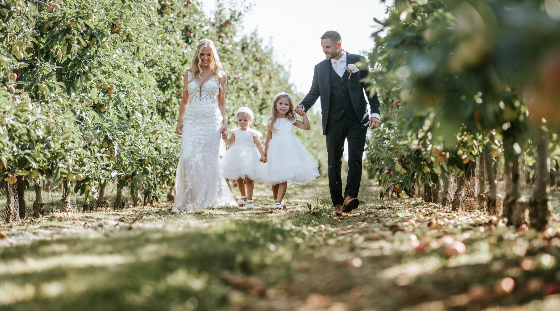 A bride and groom walking through the apple orchard with their two flower girls at The BarnYard in Kent
