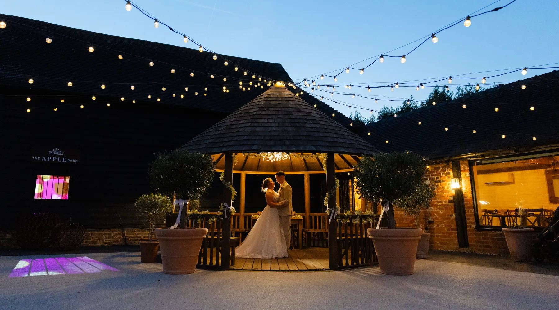 A couple beneath the Courtyard gazebo at dusk, festoon lights strung across the sky at The BarnYard