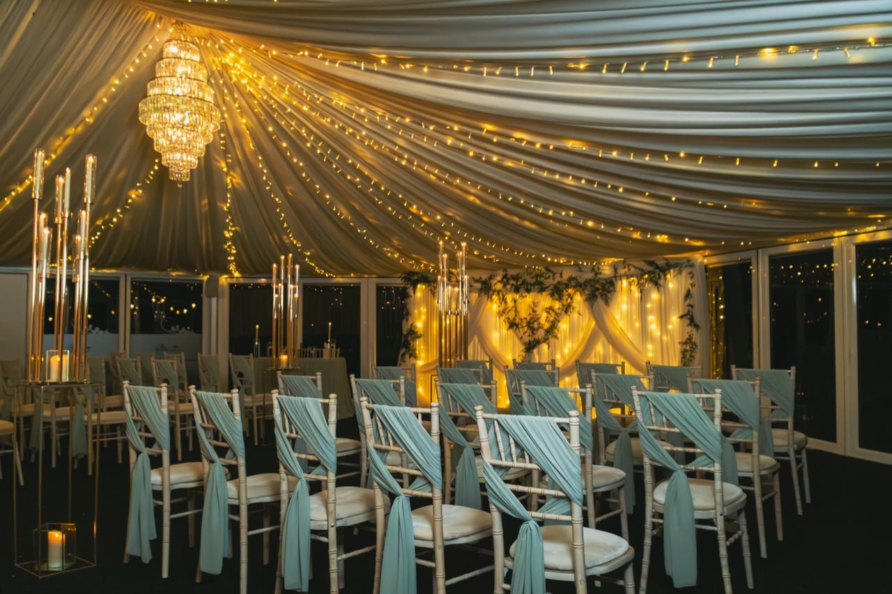 Inside the Arboretum glass pavilion at The BarnYard, a crystal chandelier and fairy-lit ceiling above chairs set for a ceremony